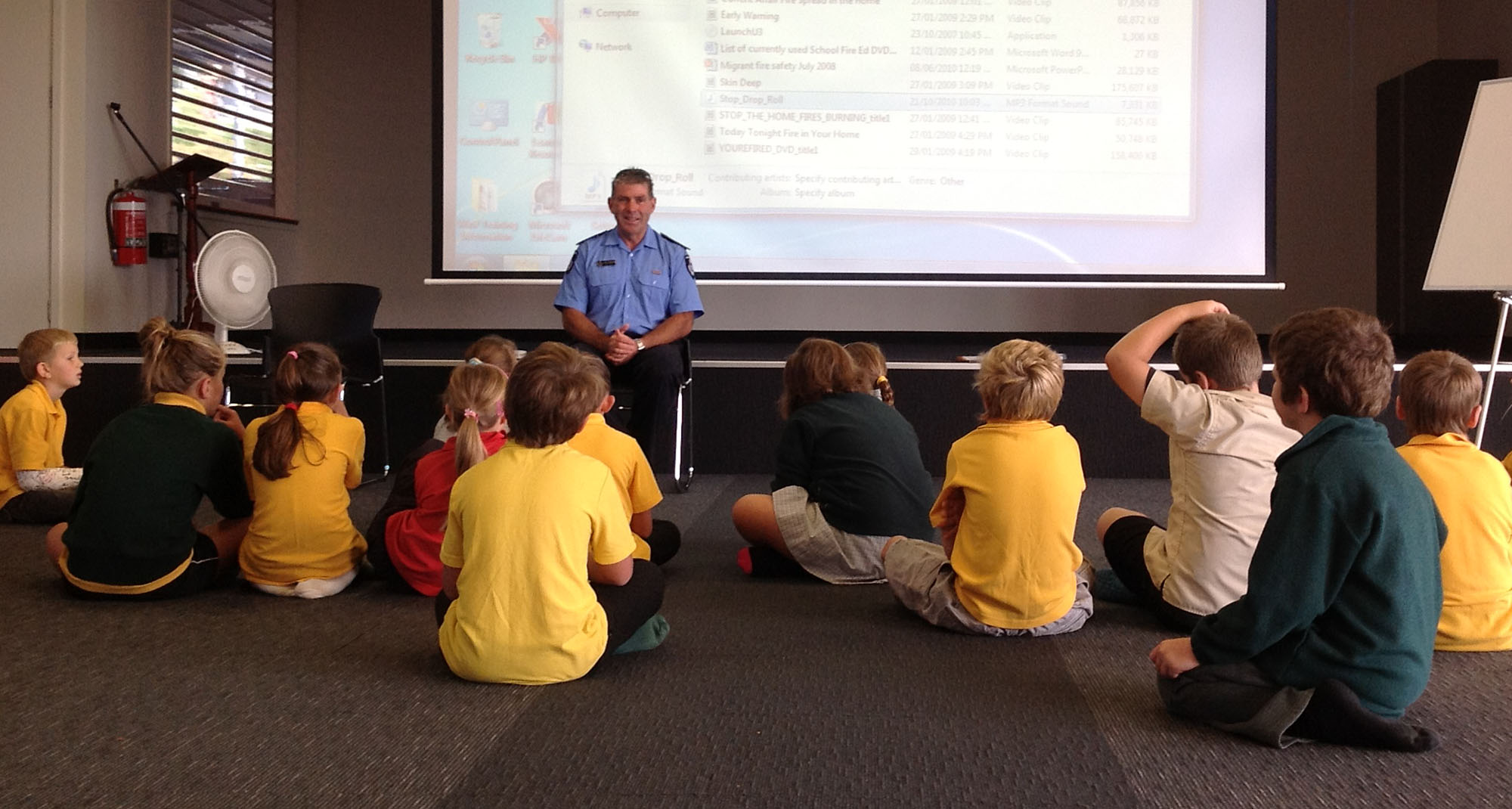 A firefighter is sitting in classroom giving fire safety tips to children sitting in front of him on the floor. A projector screen is seen behind him.