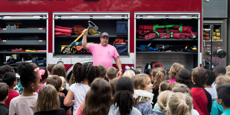 A firefighter is teaching safety tips to school children. Behind him safety equipment are arranged in shelves.